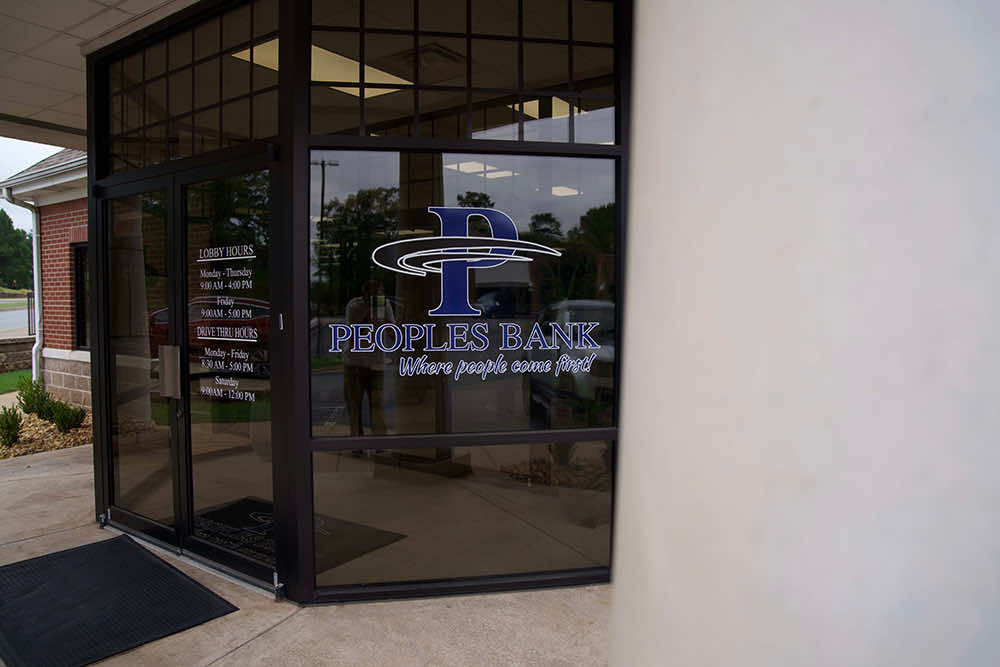 Entrance doors at a Peoples Bank branch with lobby and drive-thru hours posted on the glass