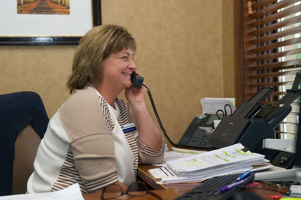 A Peoples Bank employee smiling while taking a call at a paperwork-covered desk